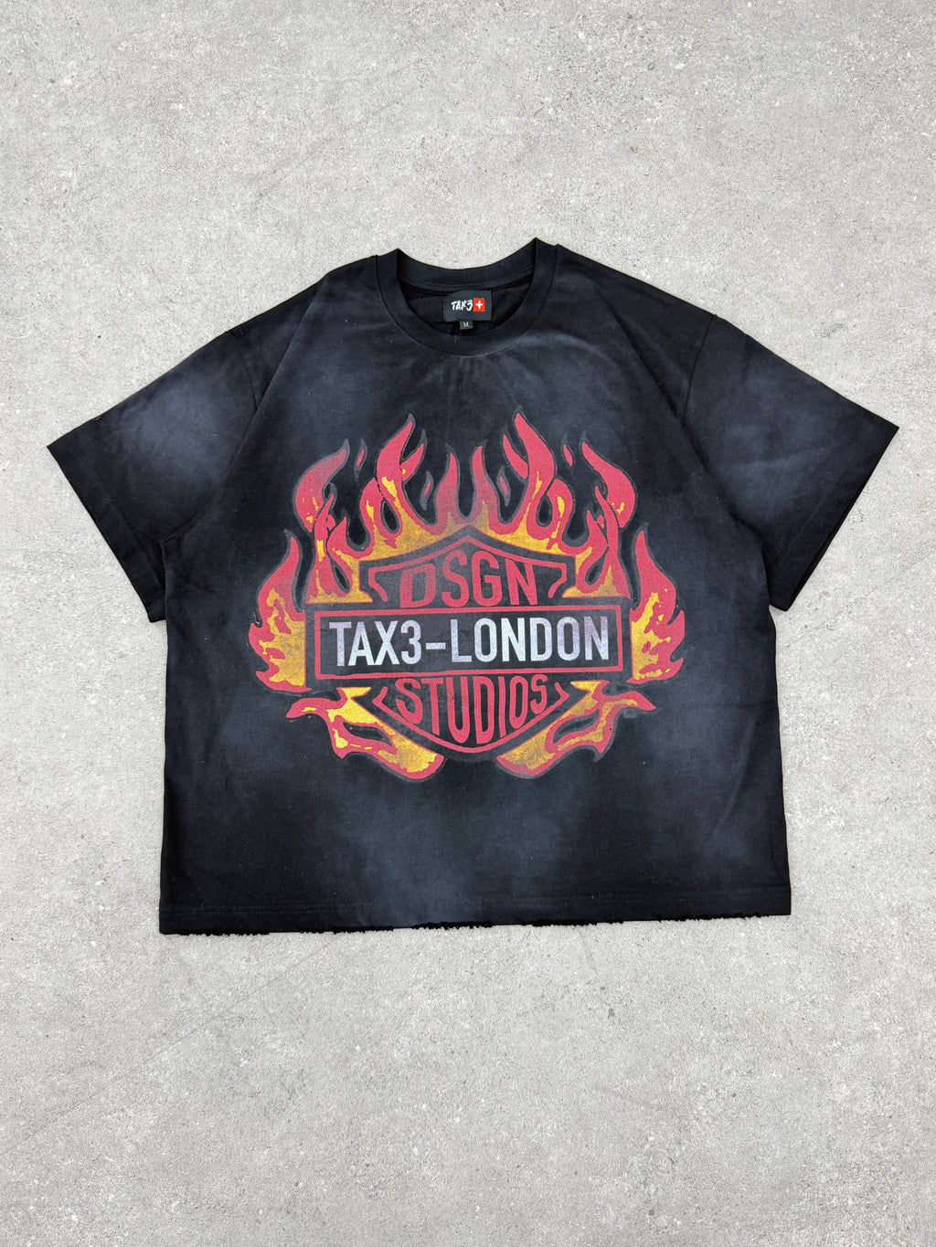 Black t-shirt with fiery graphic design and 'TAX3 LONDON STUDIOS' text on a acid wash fabric surface.